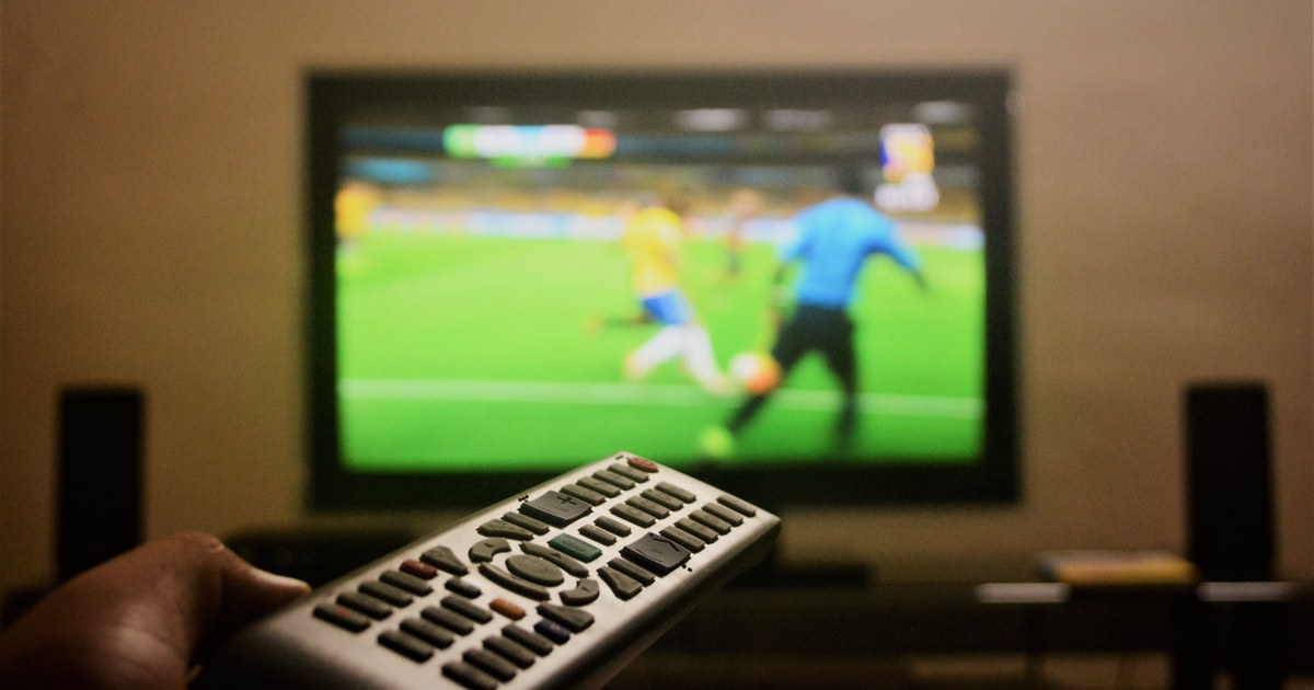 Peacock Content Acquisition Strategy: A 2026 Guide 1 Hand holding a remote control in front of a television showing a live football match, representing Peacock's sports-driven content acquisition strategy